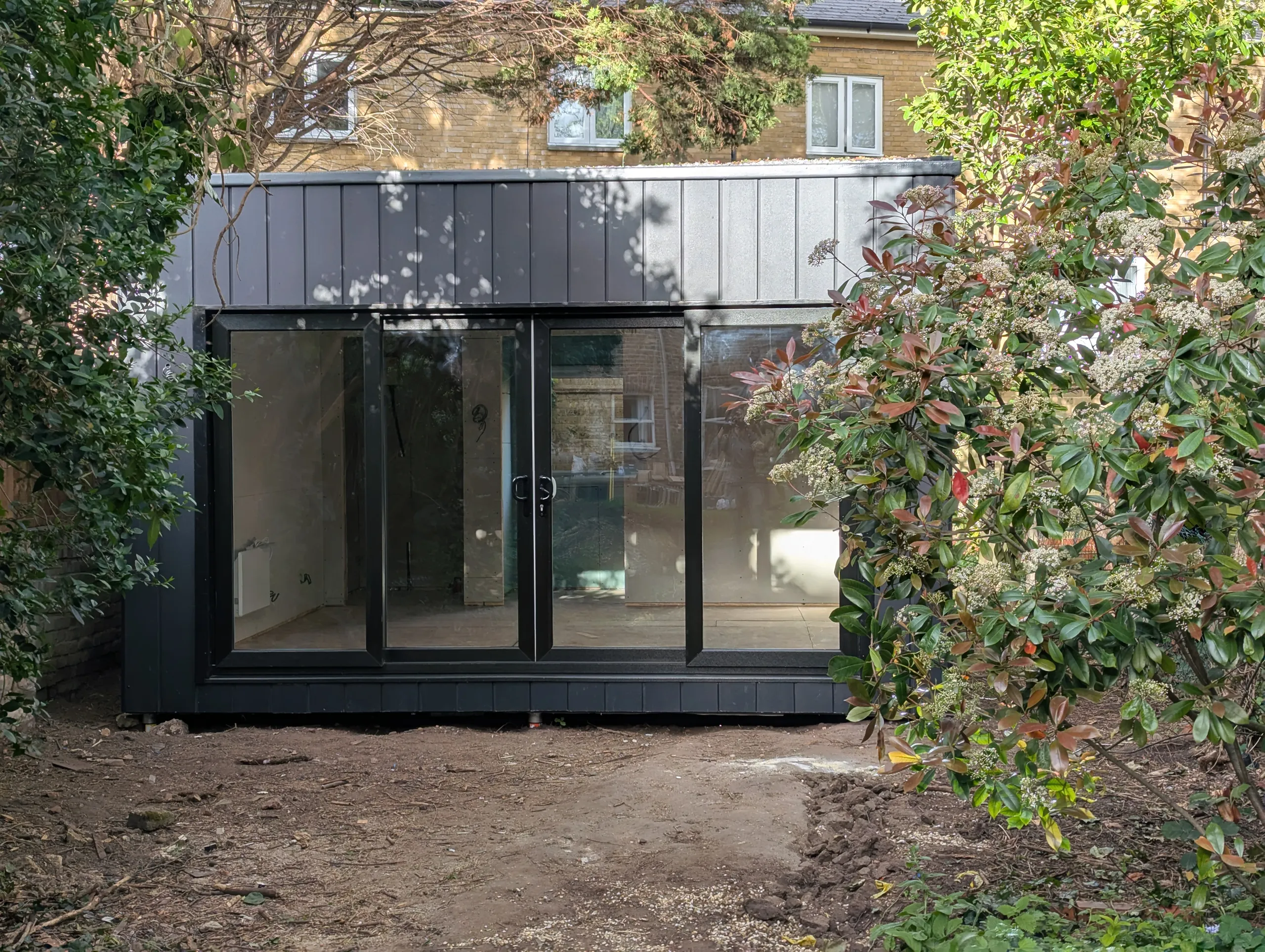 The completed garden room seen straight on, a black cement board clad cube with full width glazing and no roof overhang