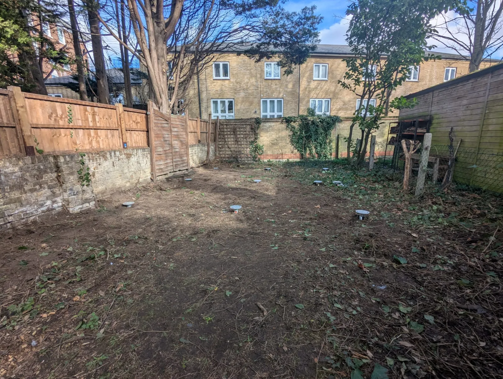 The cleared garden plot with the ground screw heads installed in position, the protected tree overhead and the neighbouring terrace beyond